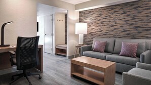 A modern hotel room with a grey sofa, a wooden coffee table, a black office chair, a standing lamp, and a patterned wall. - Sheraton Richmond Airport Hotel (Sandston)