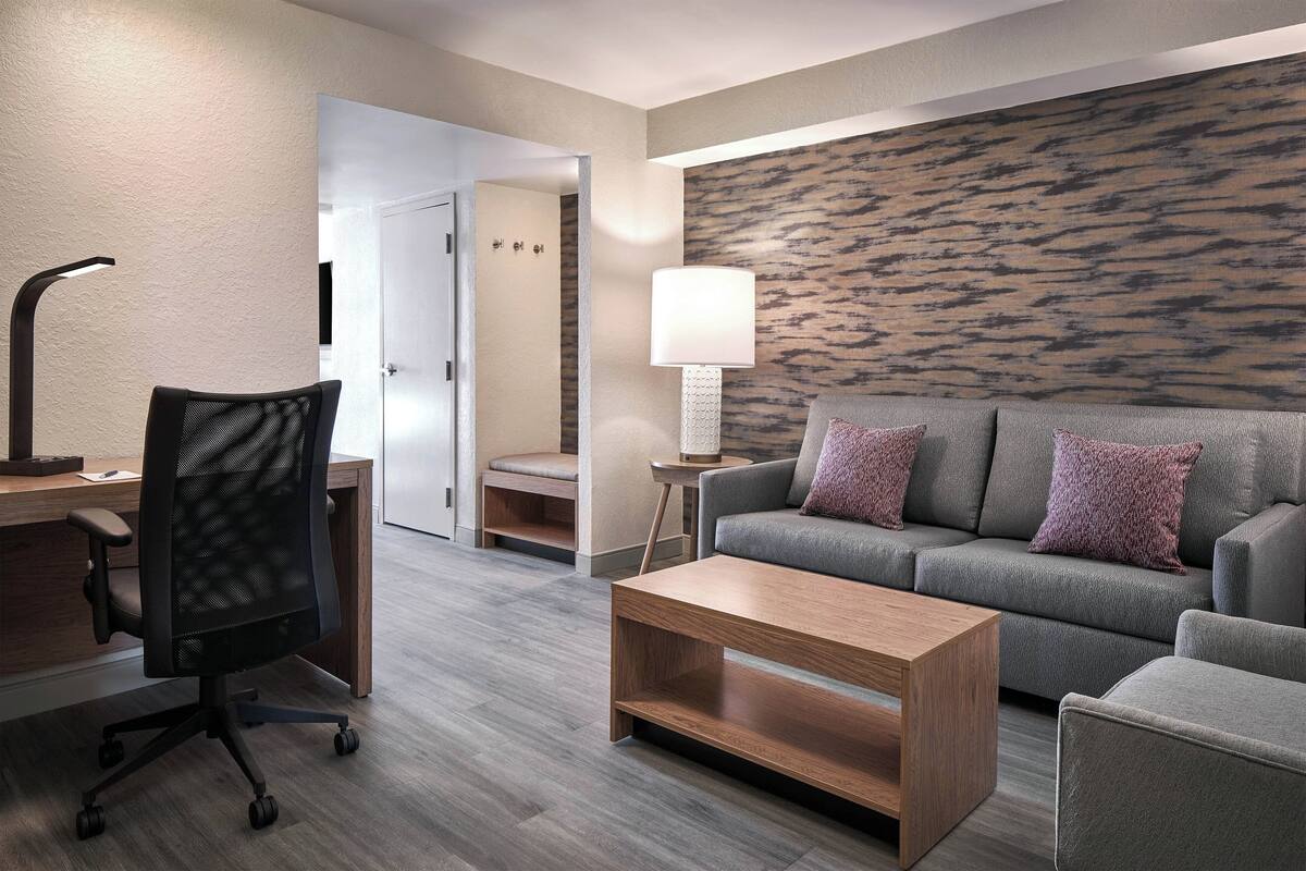 Una habitación de hotel moderna con un sofá gris, una mesa de centro de madera, una silla de oficina negra, una lámpara de pie y un papel tapiz estampado.