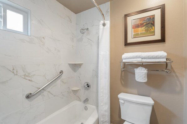 A bathroom with a bathtub, a shower, a toilet, a handrail, a window, and a towel rack with towels.