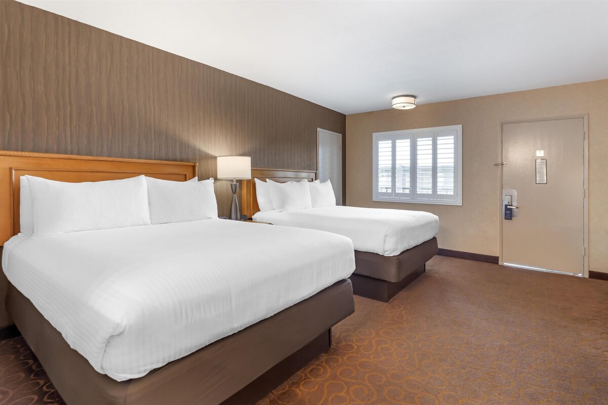 A hotel room with two beds, a wooden headboard, a nightstand with a lamp, a window with blinds, and a door with a lock.