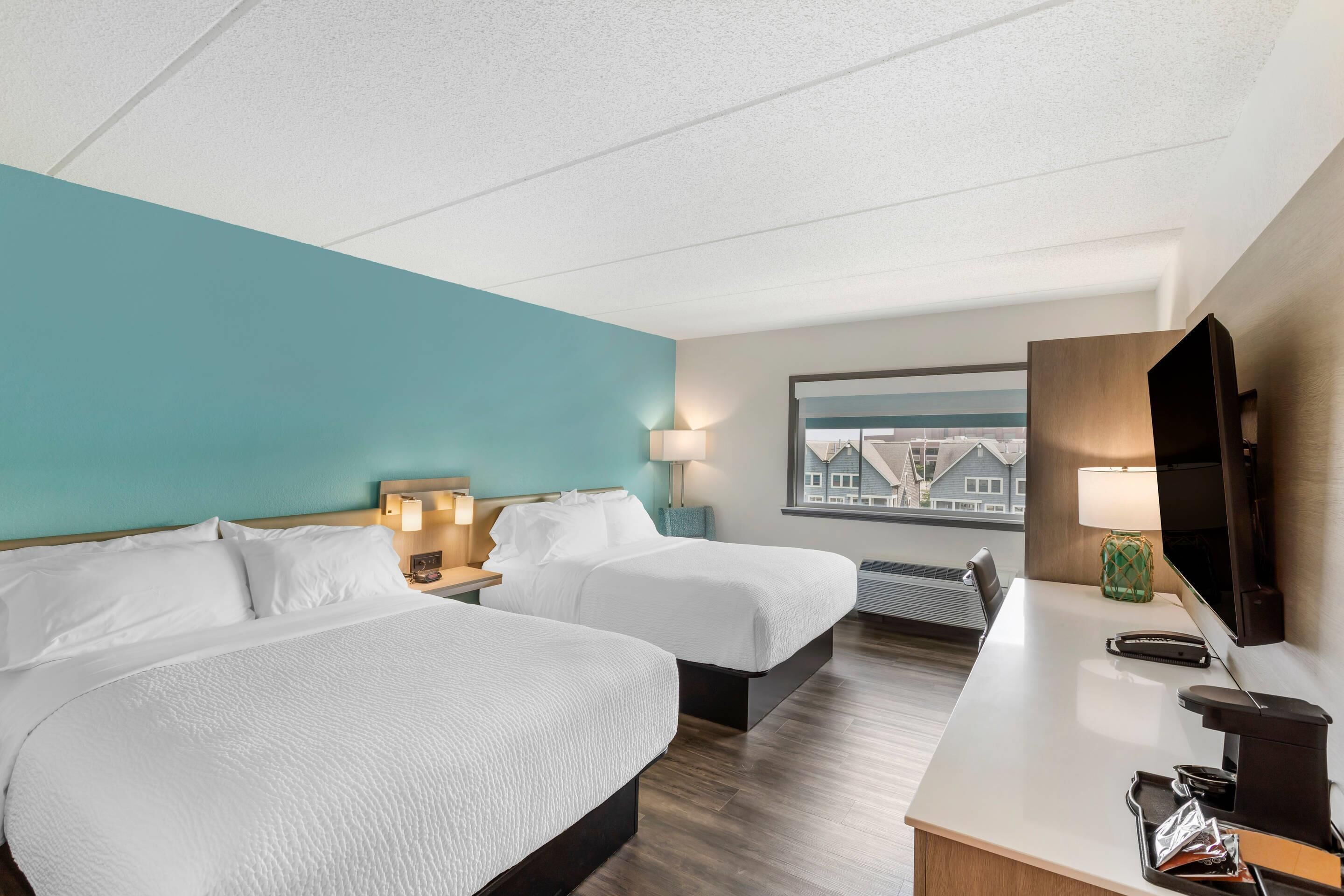A hotel room with two beds, a desk, a TV, and a window with a view of buildings.