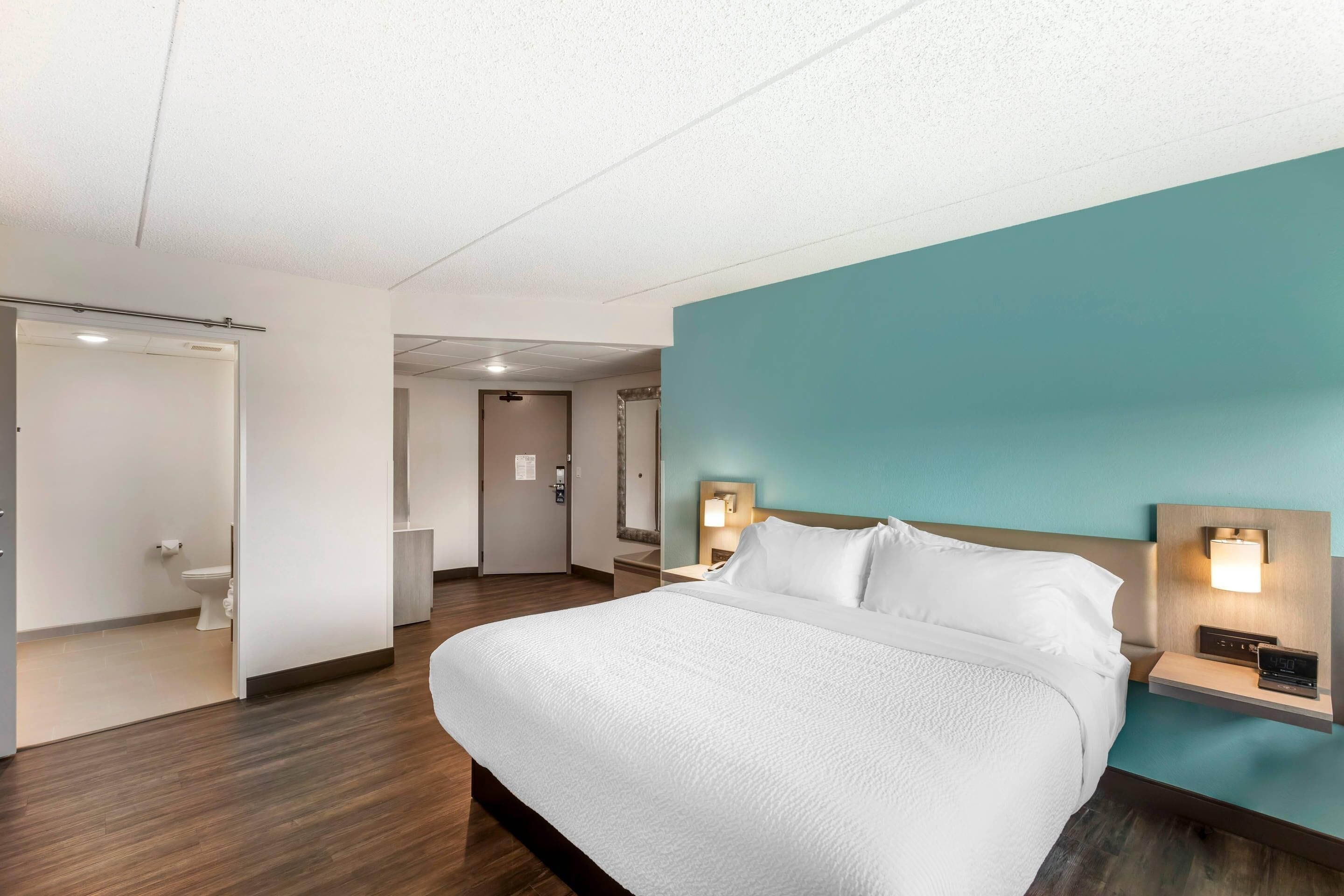 Una habitación de hotel con cama, mesita de noche con lámpara, baño con inodoro y puerta que da a otra habitación.