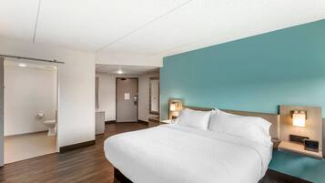 Una habitación de hotel con cama, mesita de noche con lámpara, baño con inodoro y puerta que da a otra habitación.