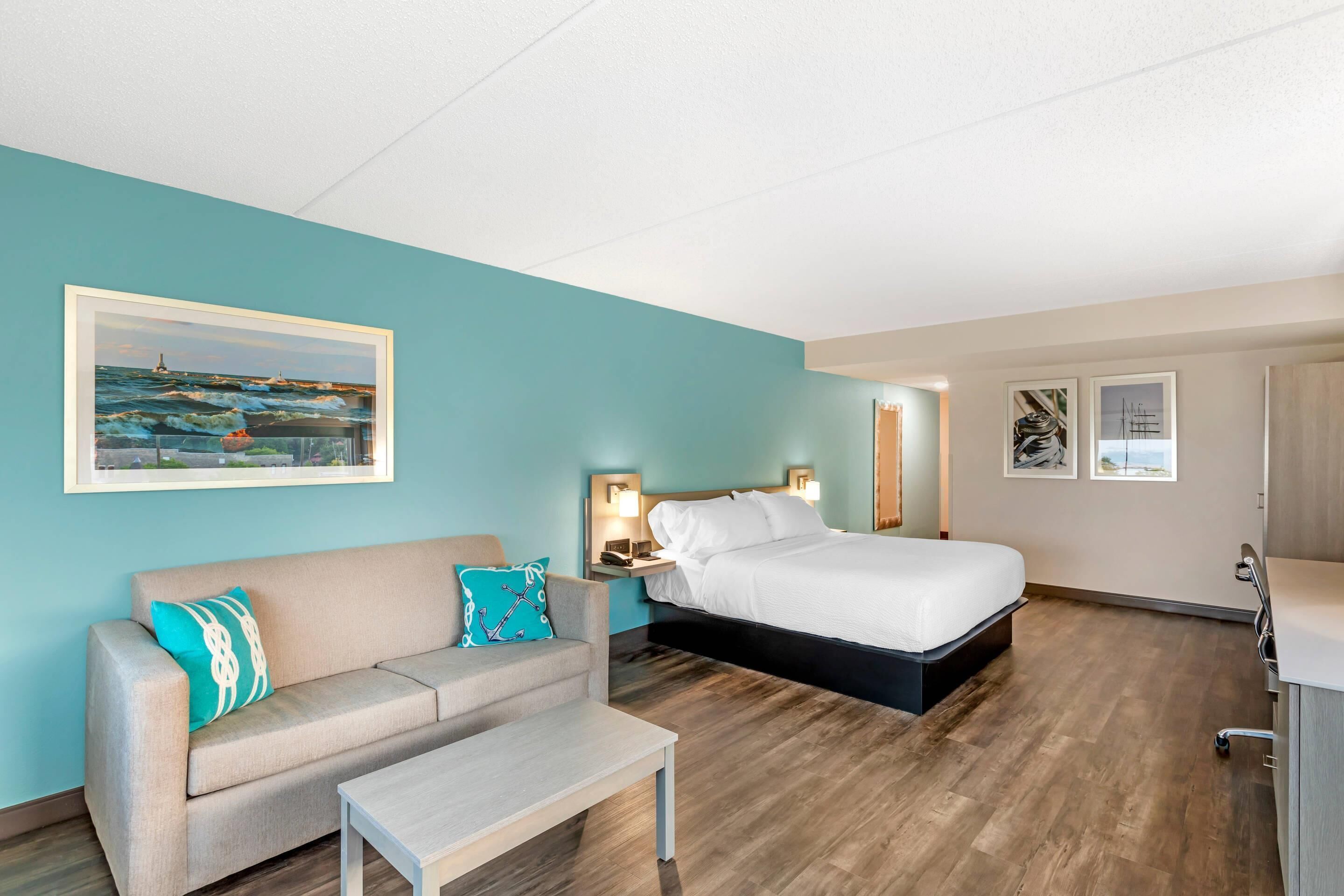 Una habitación de hotel con una cama, un sofá, una mesita de noche y un cuadro de una escena costera.