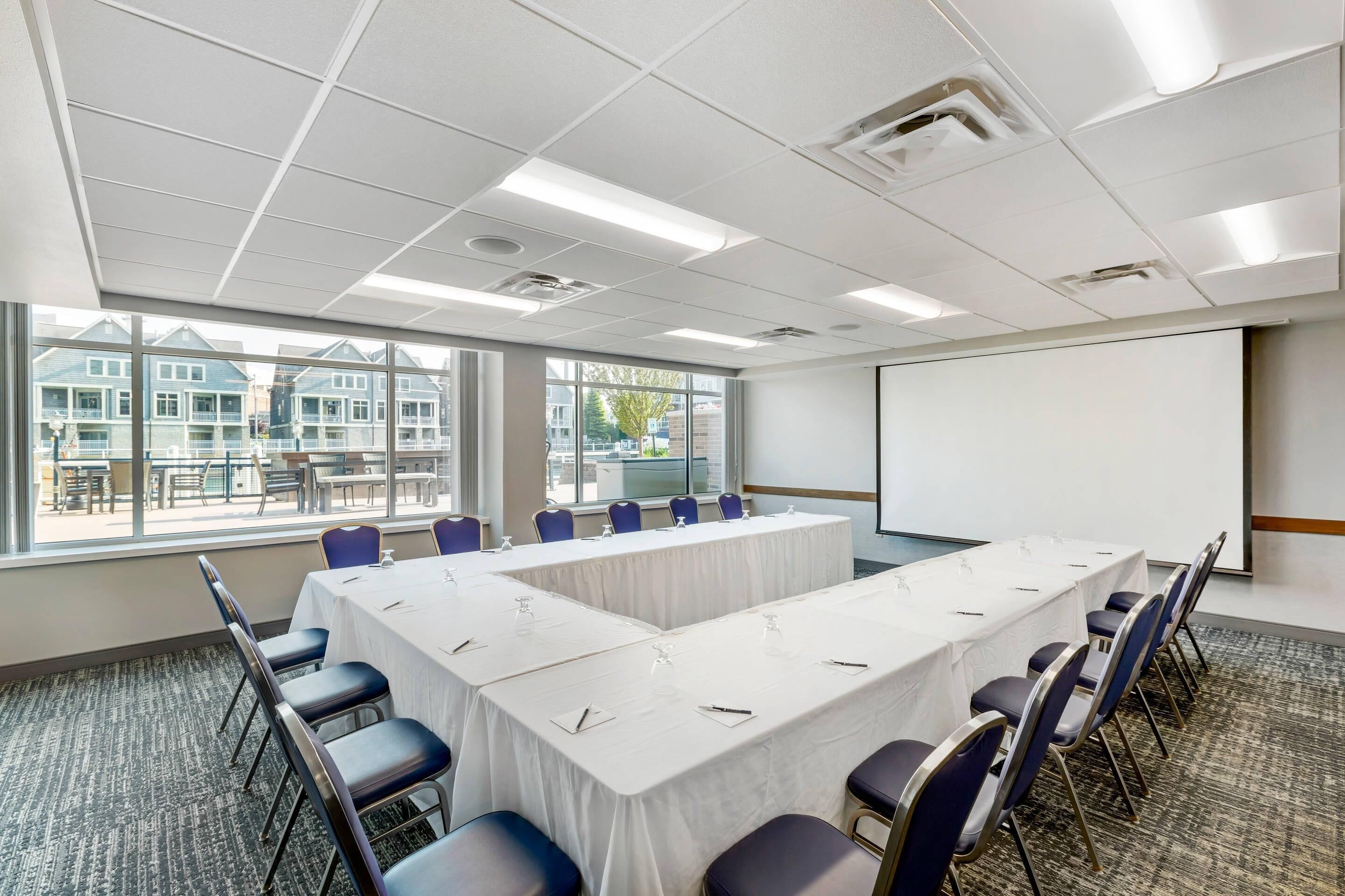 A conference room with a long table, chairs, a projector screen, and large windows with a view of buildings.