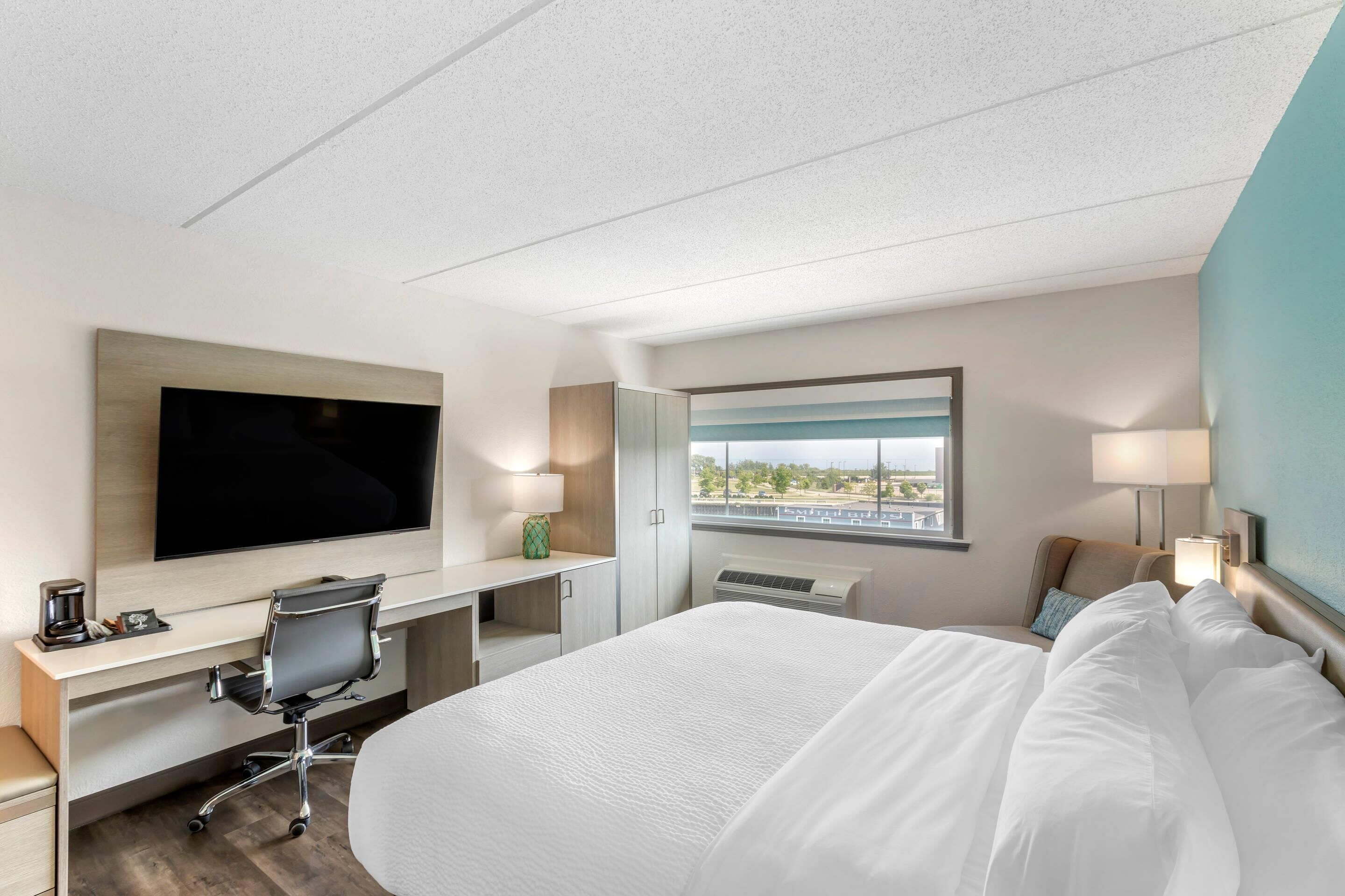 A modern hotel room with a large bed, a desk with a flat-screen TV, a comfortable chair, a window with a city view, and a bedside table with a lamp.