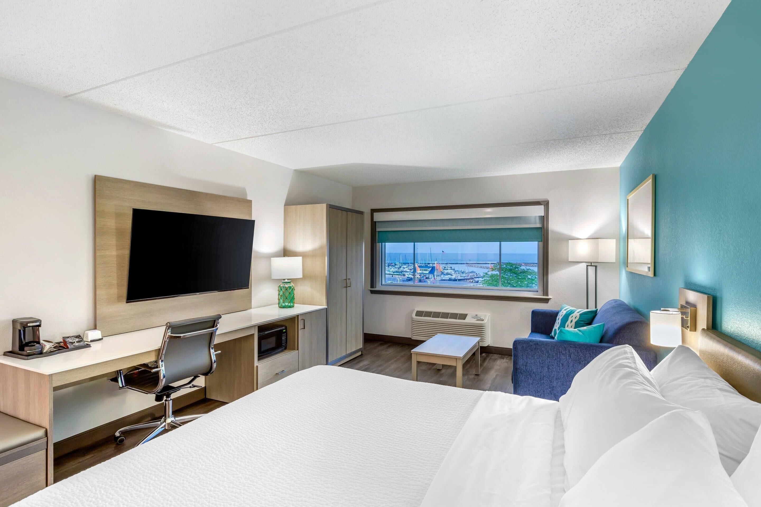 A hotel room with a bed, desk, chair, TV, window with a view, and a small table.