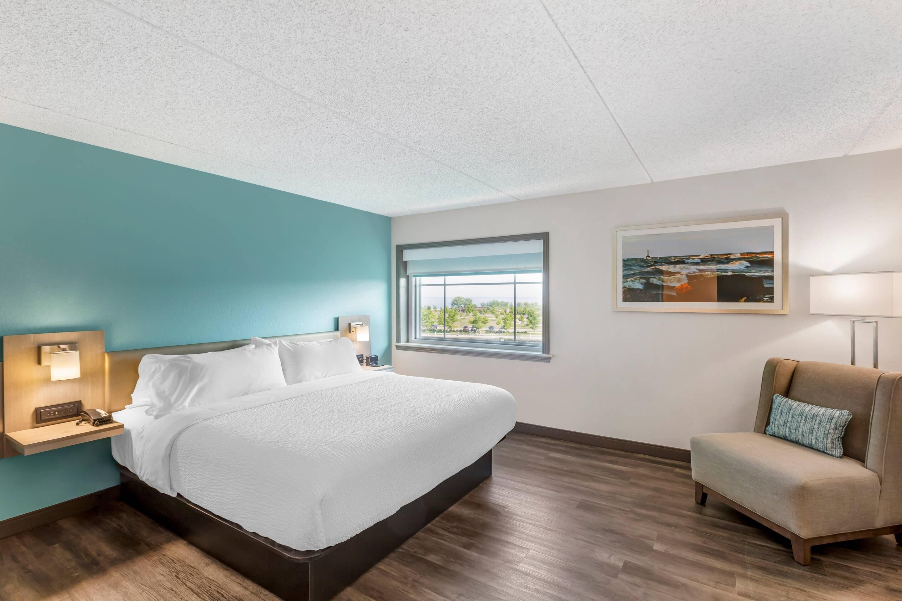 A hotel room with a bed, a nightstand, a lamp, a window with a view, a chair, and a framed picture on the wall.