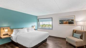 A hotel room with a bed, a nightstand, a lamp, a window with a view, a chair, and a framed picture on the wall.