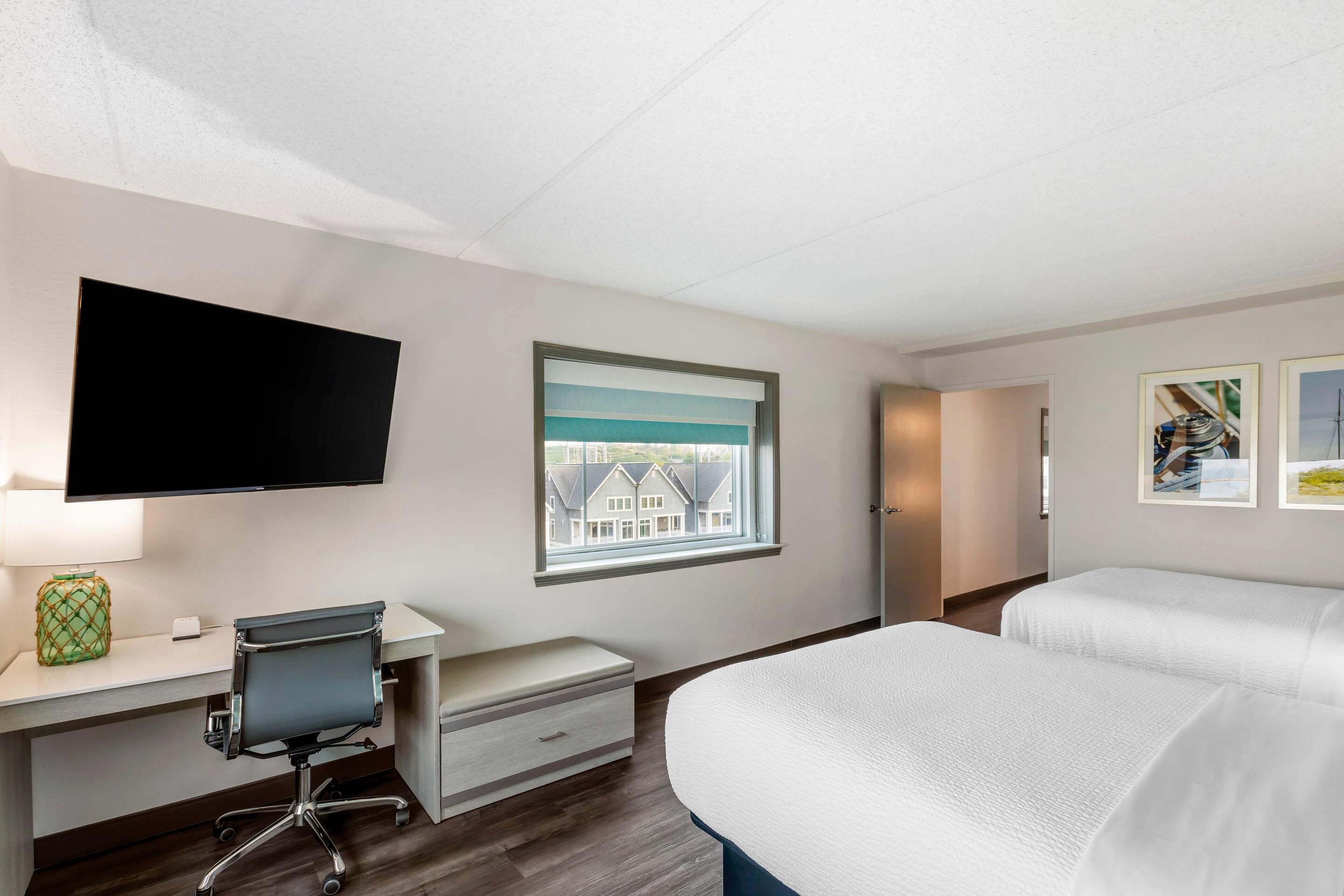 A hotel room with a bed, a desk with a chair, a television, a window with a view of houses, and framed pictures on the wall.