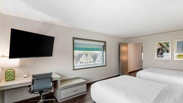 A hotel room with a bed, a desk with a chair, a television, a window with a view of houses, and framed pictures on the wall.
