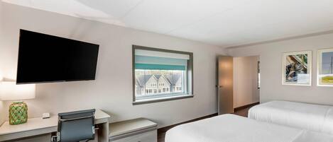 A hotel room with a bed, a desk with a chair, a television, a window with a view of houses, and framed pictures on the wall.
