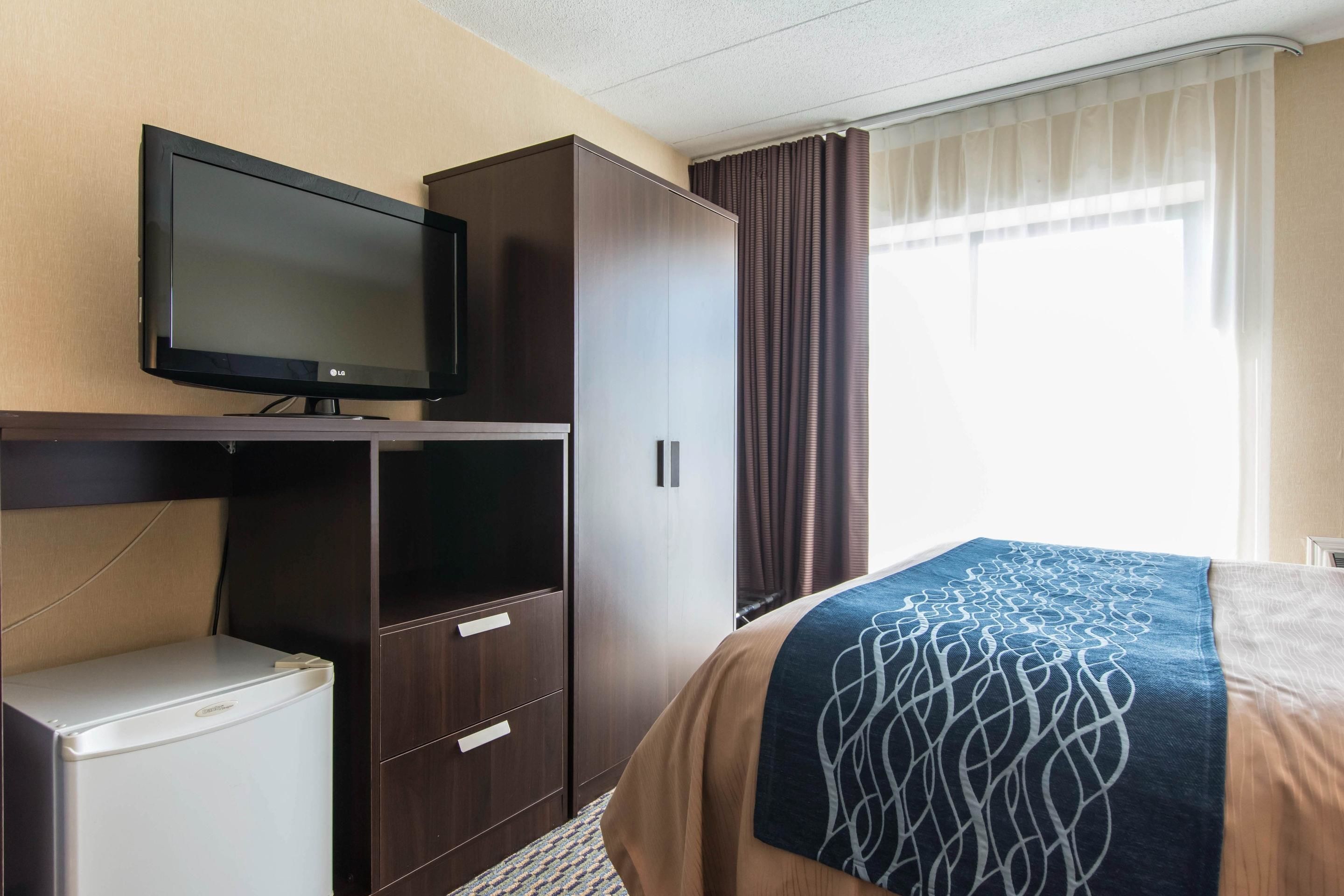 A hotel room with a bed, a television on a stand, a wardrobe, and a small refrigerator.