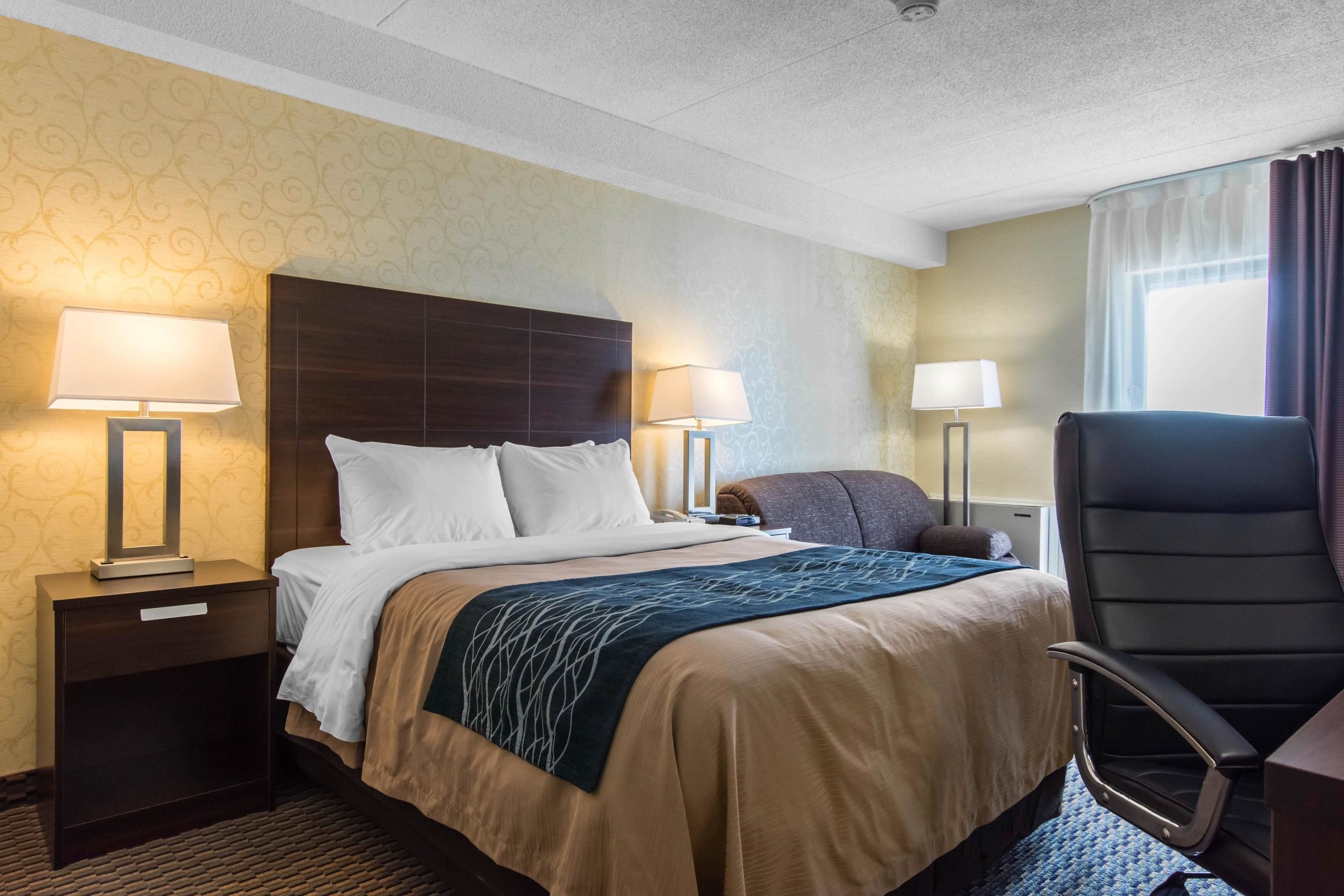 A hotel room with a large bed, two bedside lamps, a dark headboard, a desk with a chair, and a window with curtains.