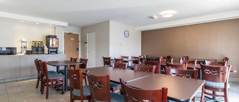 Un comedor con varias mesas y sillas de madera, una zona de cocina con electrodomésticos, y un reloj en la pared.