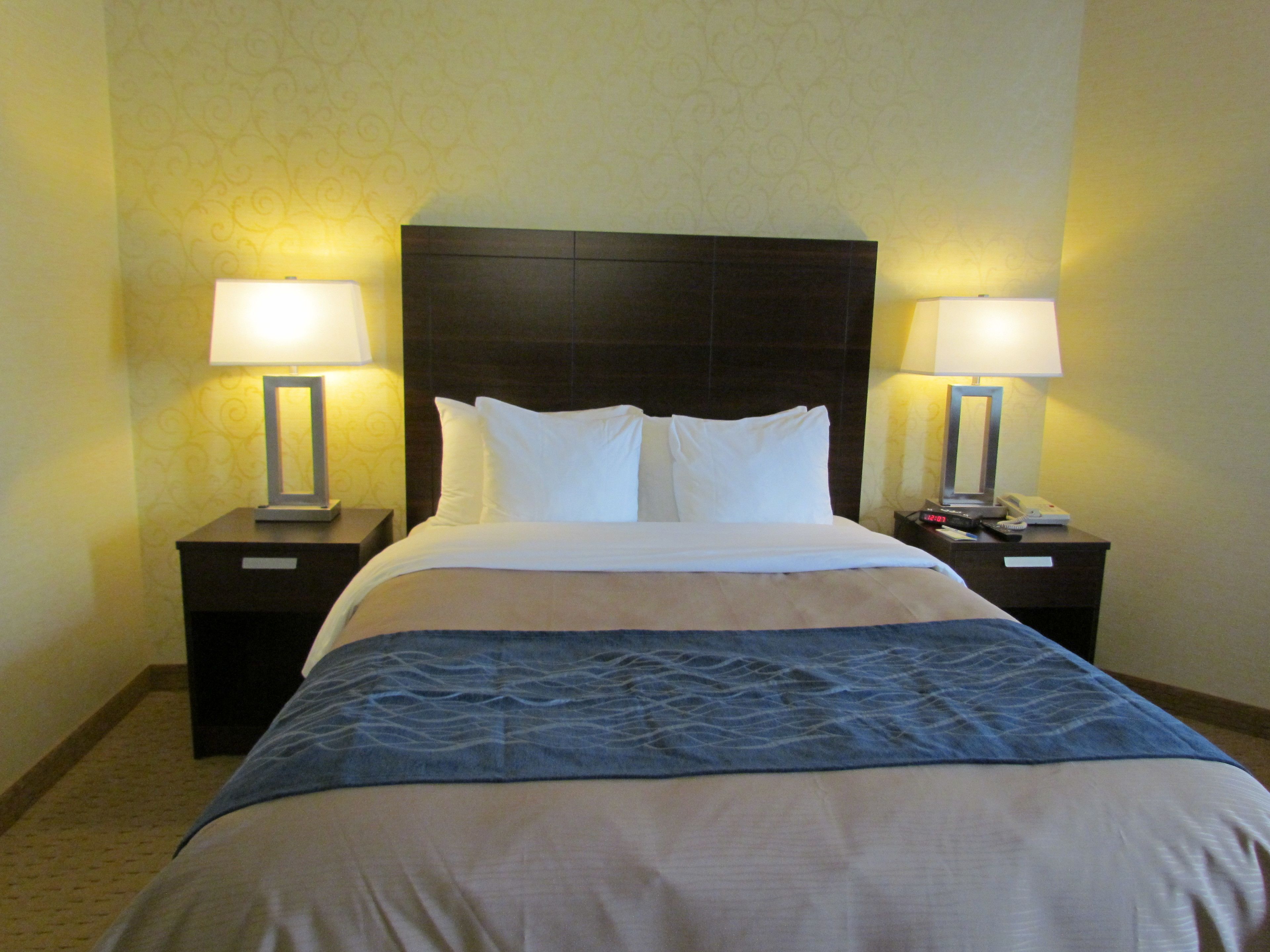 A hotel room with a large bed, two bedside tables with lamps, a nightstand with a phone and remote control, and a patterned wallpaper.