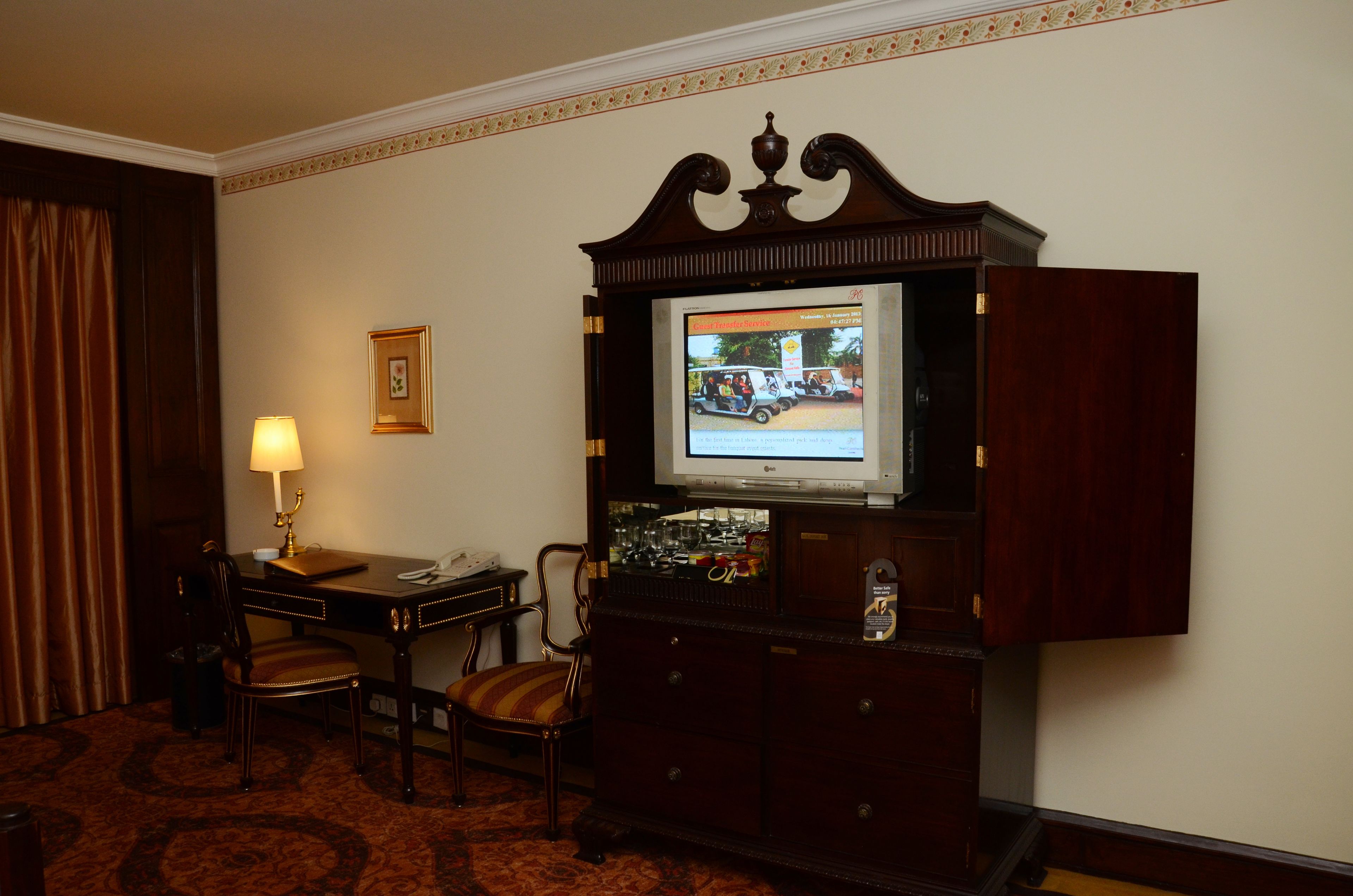 Een hotelkamer met een televisie op een houten kast, een bureau met een lamp, twee stoelen en een vloerkleed.