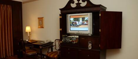 A hotel room with a television on a wooden cabinet, a desk with a lamp, two chairs, and a carpeted floor.