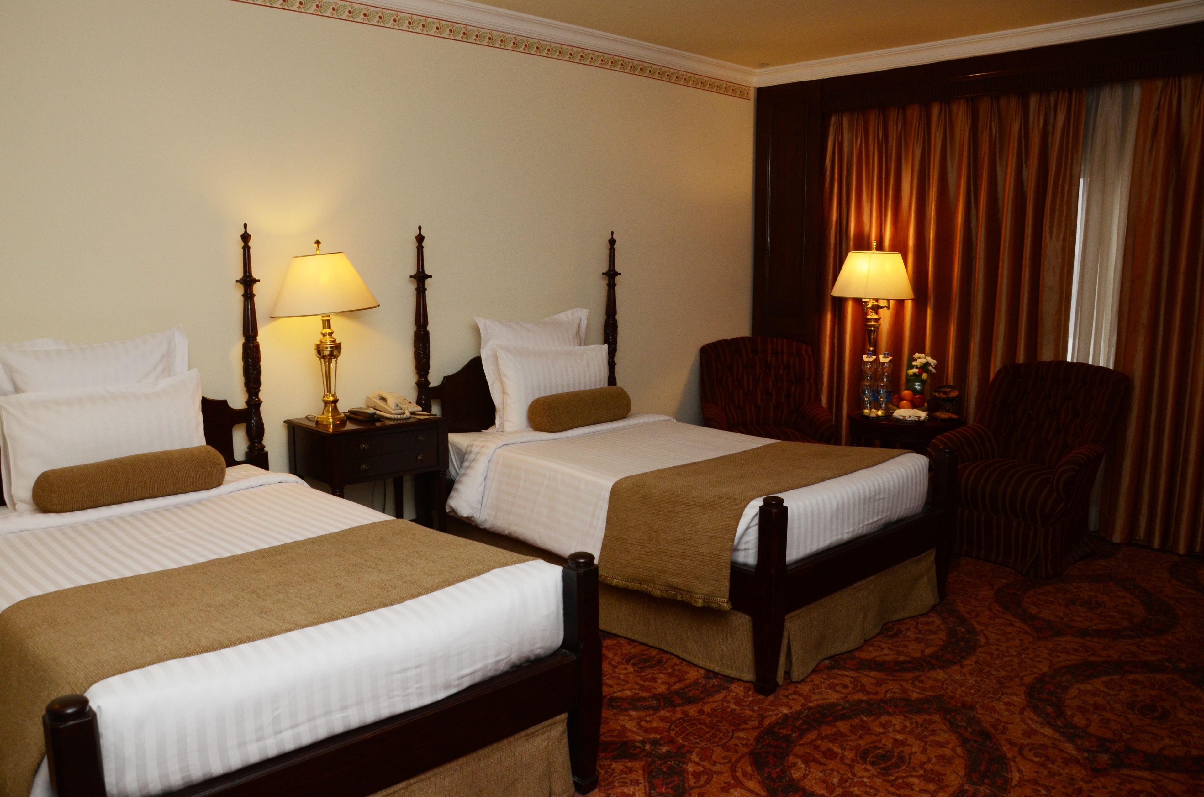 A hotel room with two beds, each with a wooden frame and white bedding. There are two bedside tables with lamps, a chair, and a large window with curtains.