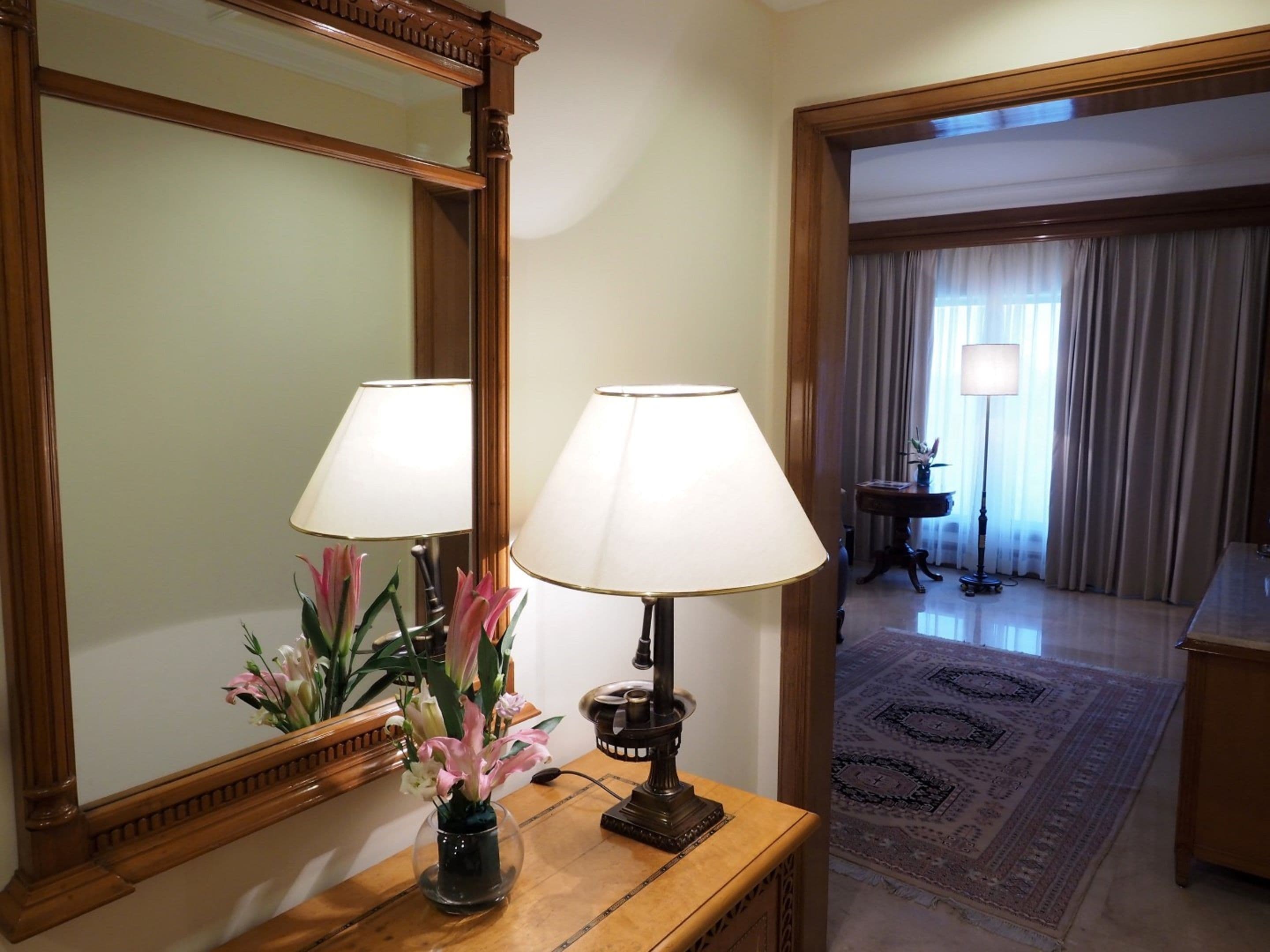 Habitación de hotel con una mesita de noche de madera, dos lámparas, un florero con flores, un espejo grande y vista a otra habitación con lámpara y cortinas.