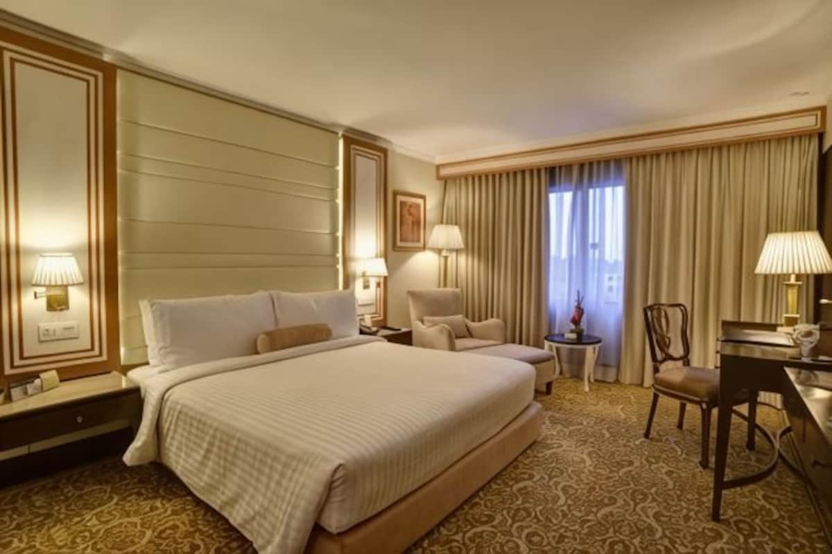 A hotel room with a large bed, two bedside tables with lamps, a desk with a chair, and a view of the outdoors through curtains.
