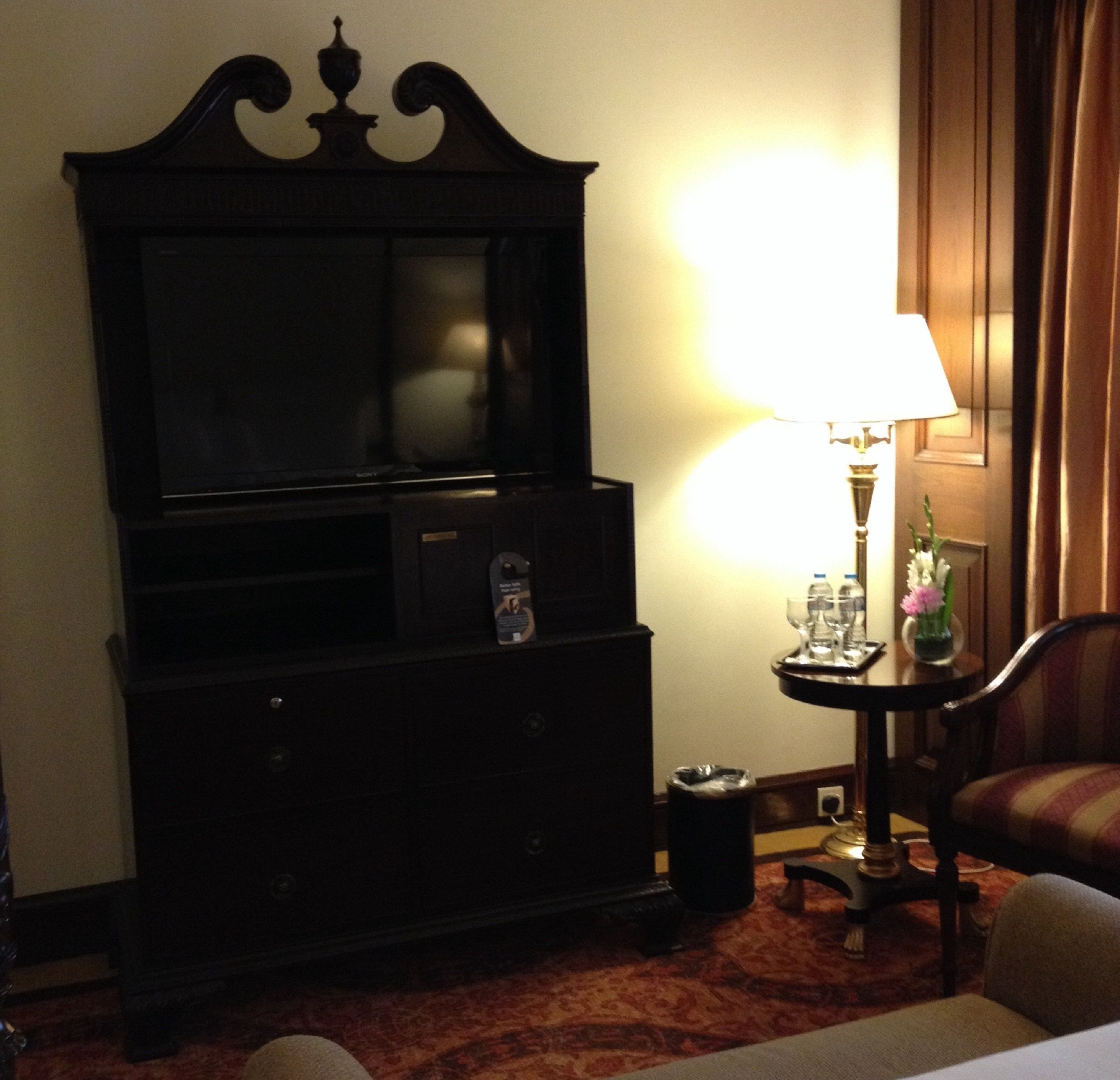 A hotel room with a large flat-screen TV, a lamp, a small table with a vase of flowers, and a chair.