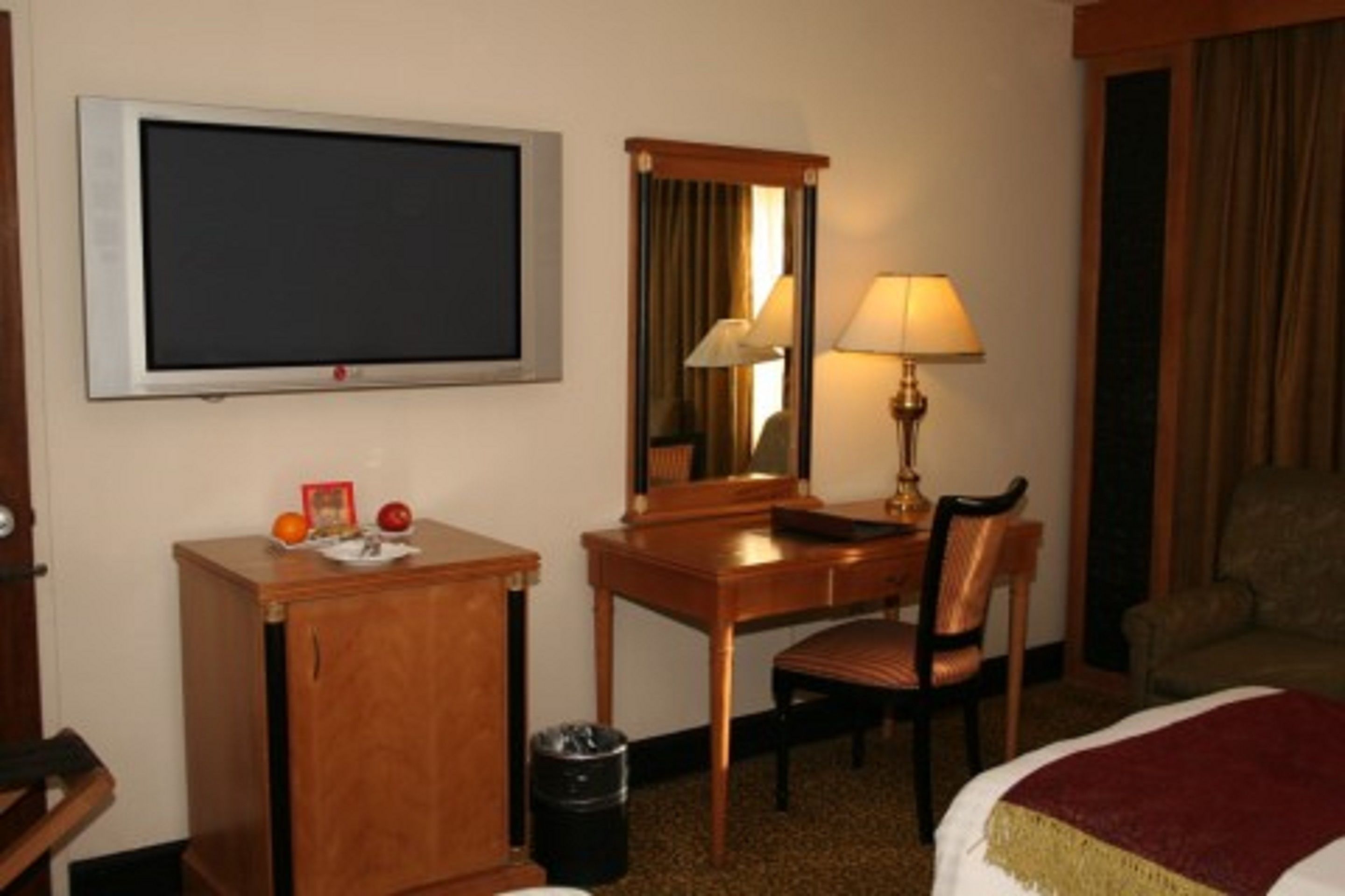 Habitación de hotel con cama, escritorio, silla, televisor, espejo, lámpara y basurero.