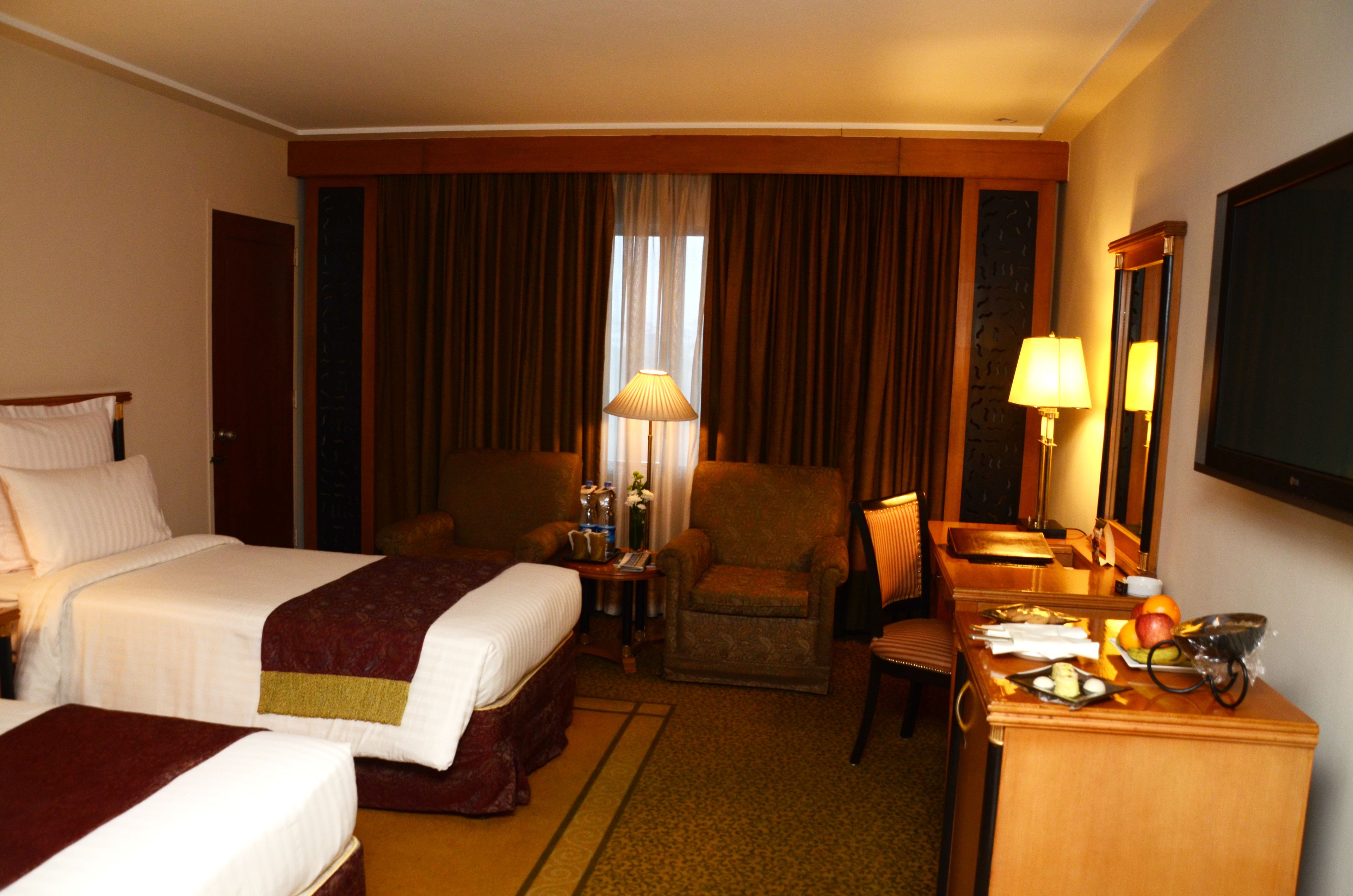 A hotel room with two beds, a desk, a chair, and a lamp.