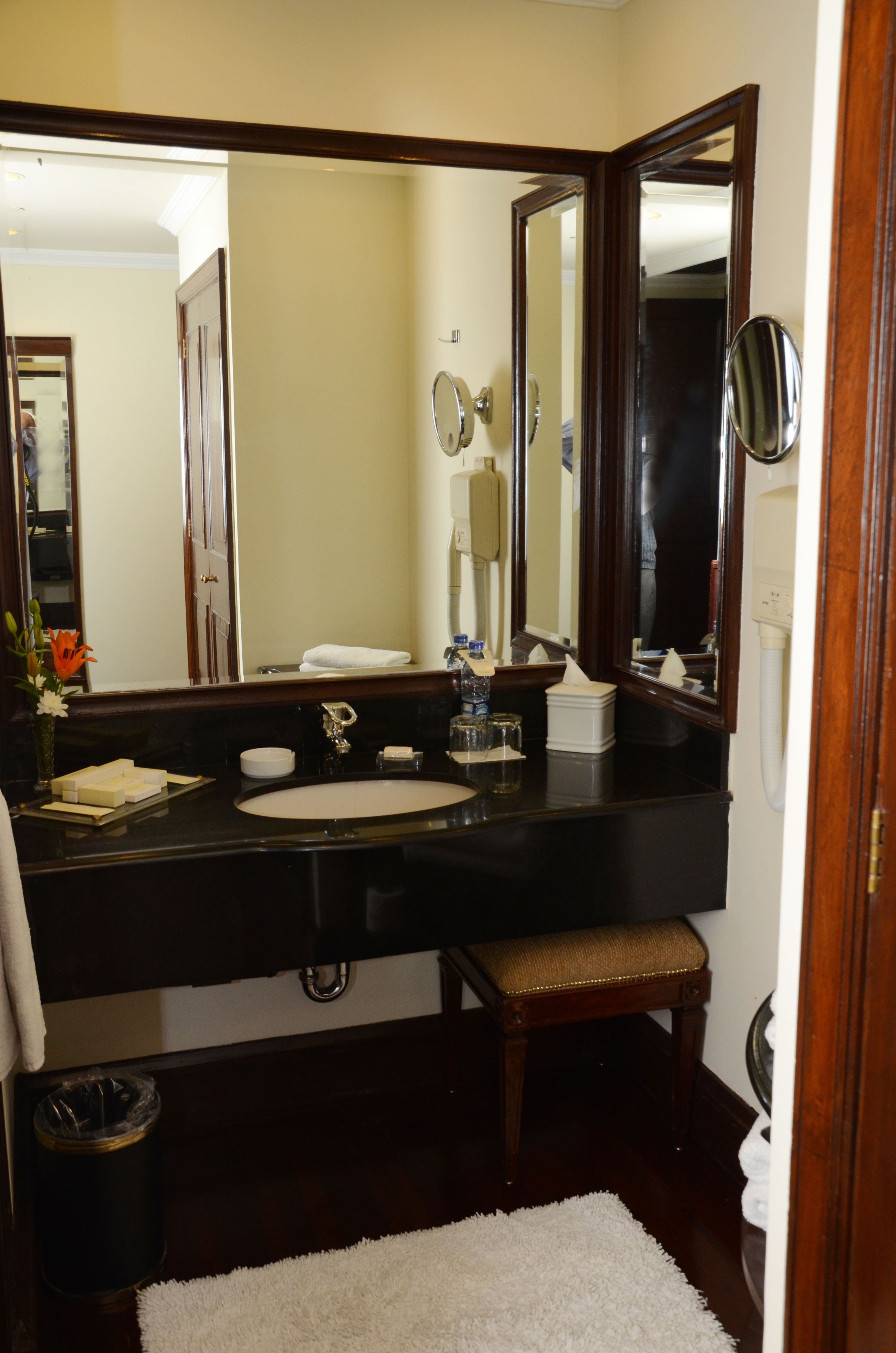 Un baño con un espejo grande, un lavabo, tocador, toallero y un cubo de basura.