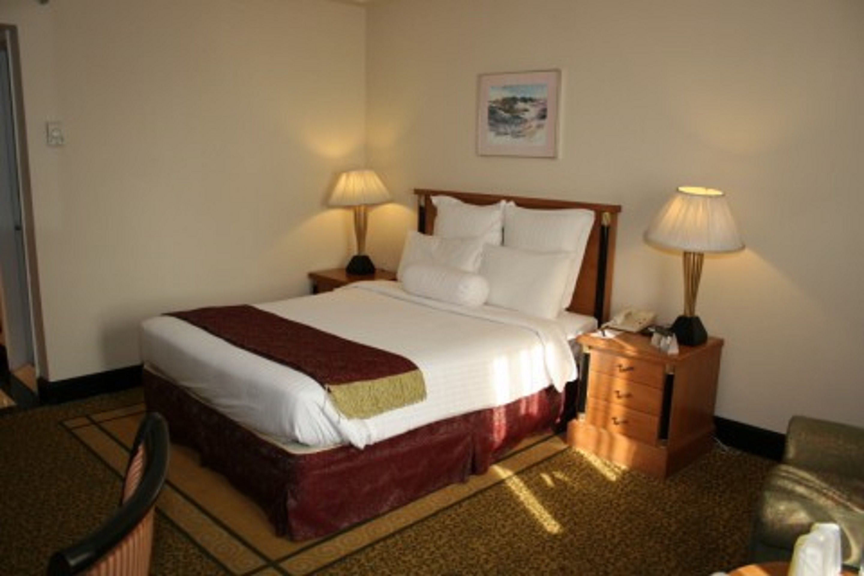 A hotel room with a large bed, two bedside tables with lamps, a wooden dresser, and a framed picture on the wall.