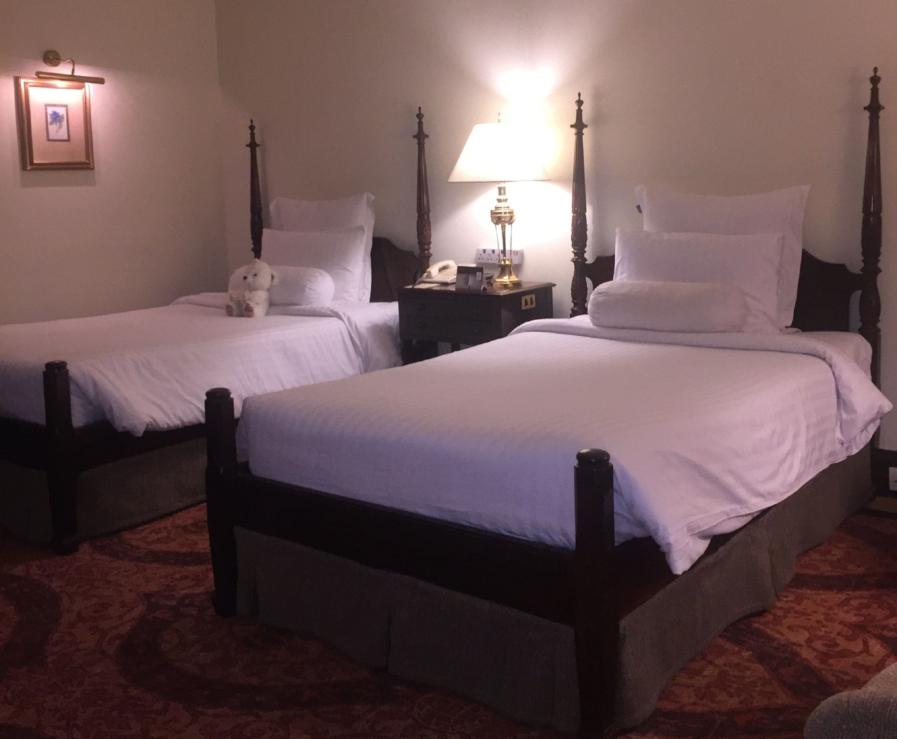 Dos camas individuales con cabeceros de madera y ropa de cama blanca, una mesita de noche con lámpara y un cuadro en la pared.