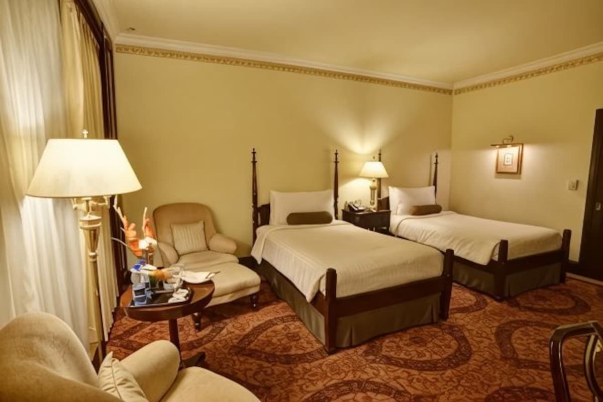 A hotel room with two beds, a chair, a table with a lamp, and a nightstand.