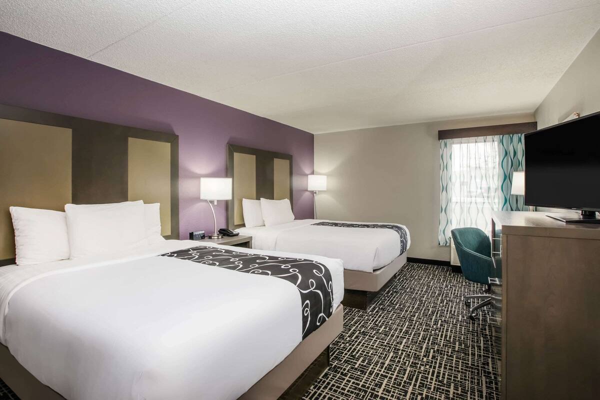 A hotel room with two beds, a desk, a television, and a window with curtains.