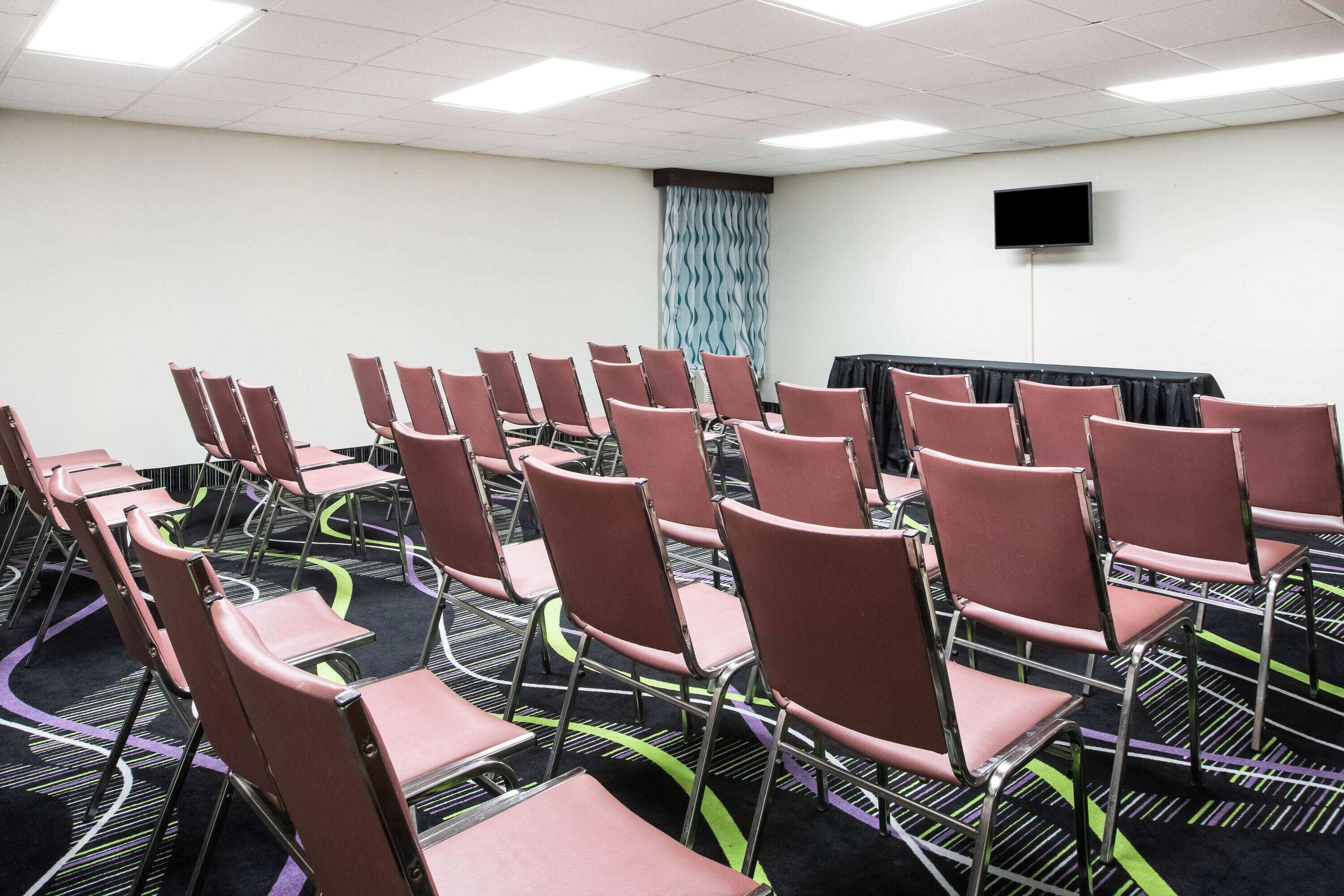 A conference room with rows of chairs, a flat-screen TV on the wall, and a patterned carpet.