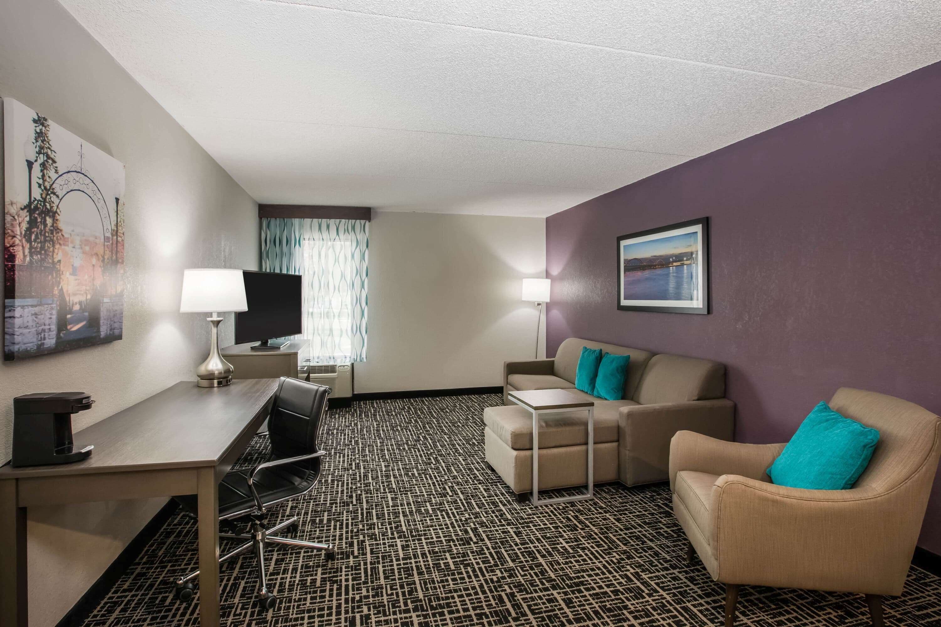 A hotel room with a desk, two chairs, a sofa, a coffee machine, a TV, and a patterned carpet.