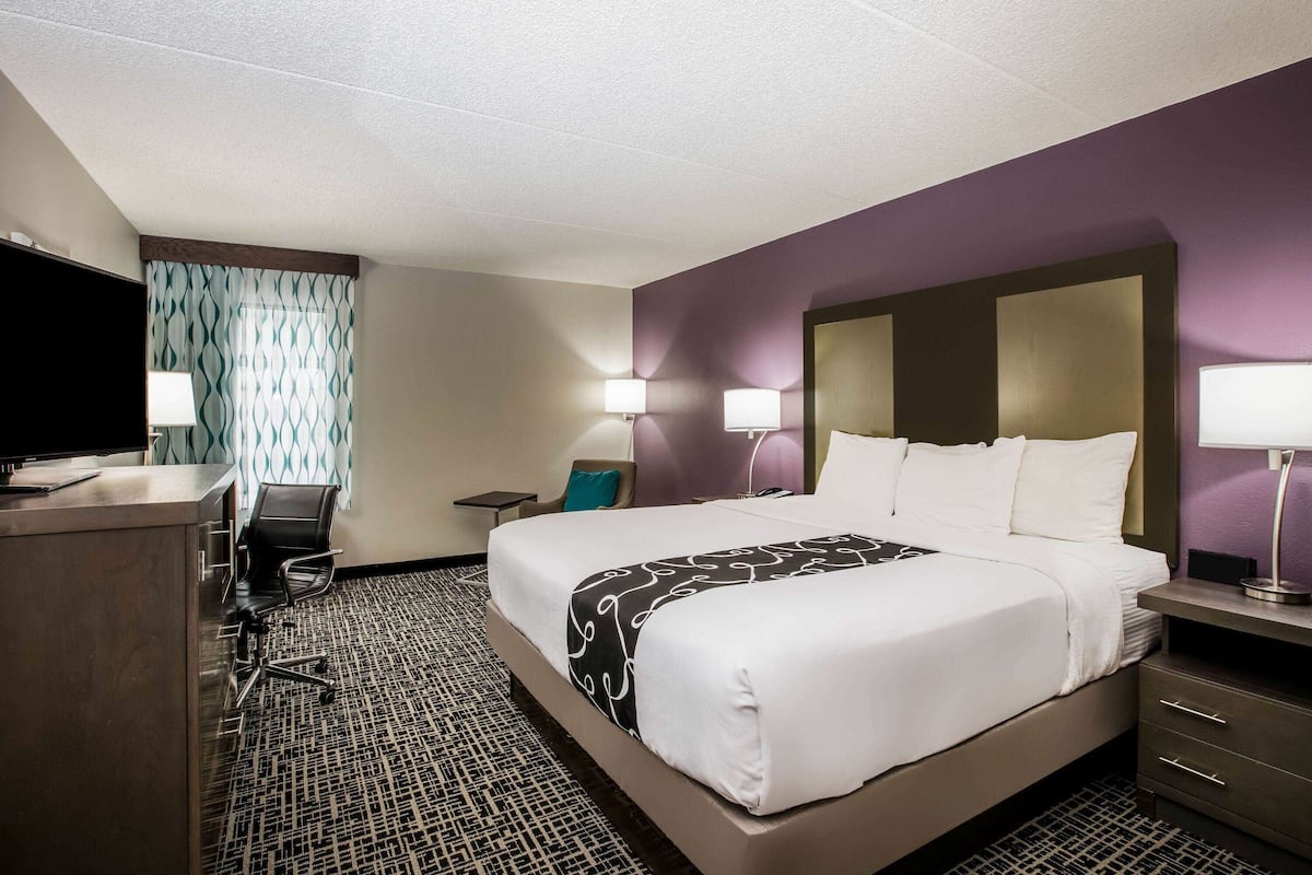 A hotel room with a large bed, a desk with a chair, a television, and a window with patterned curtains.