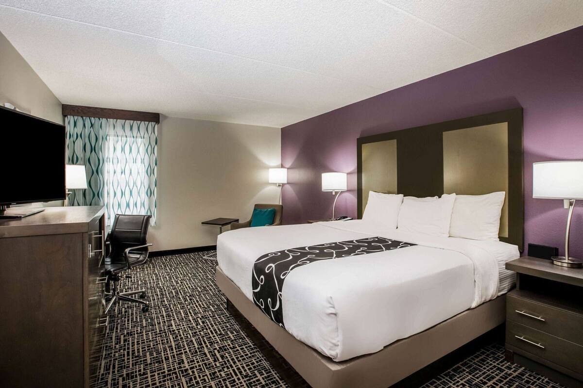 A hotel room with a bed, a desk, a chair, a TV, and a patterned rug.
