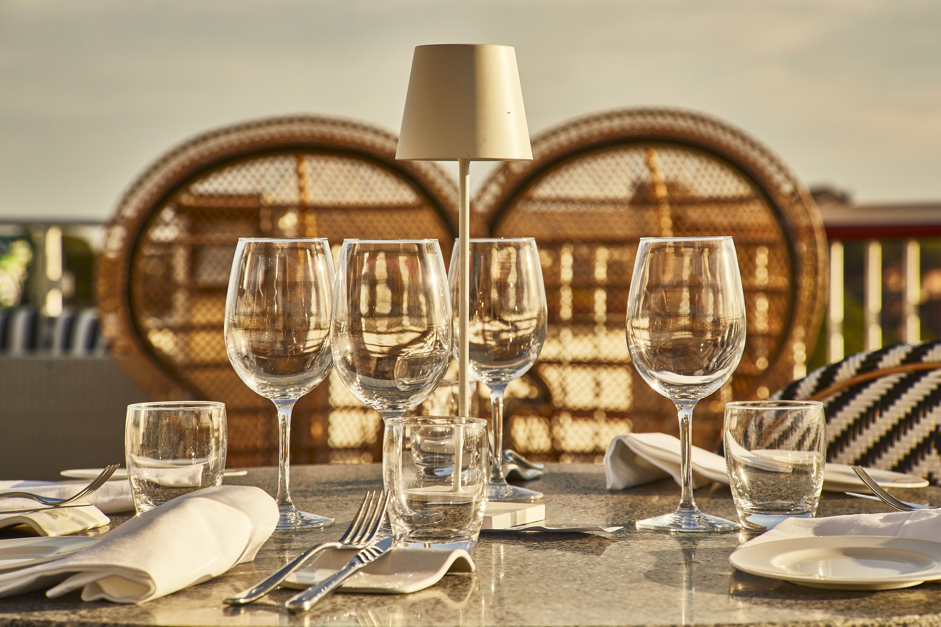 A table set for dining with multiple wine glasses, a lamp, and wicker chairs in the background.