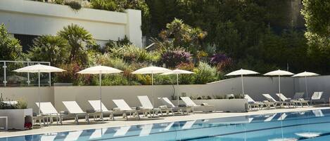 A swimming pool with lounge chairs and umbrellas, surrounded by greenery and a building in the background.