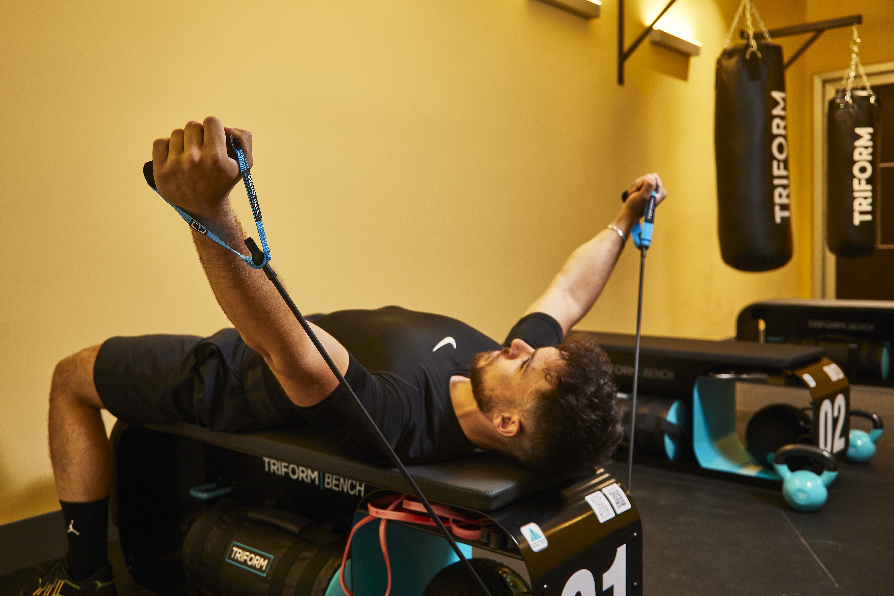 A person lying on a TRIFORM bench in a gym, using resistance bands.