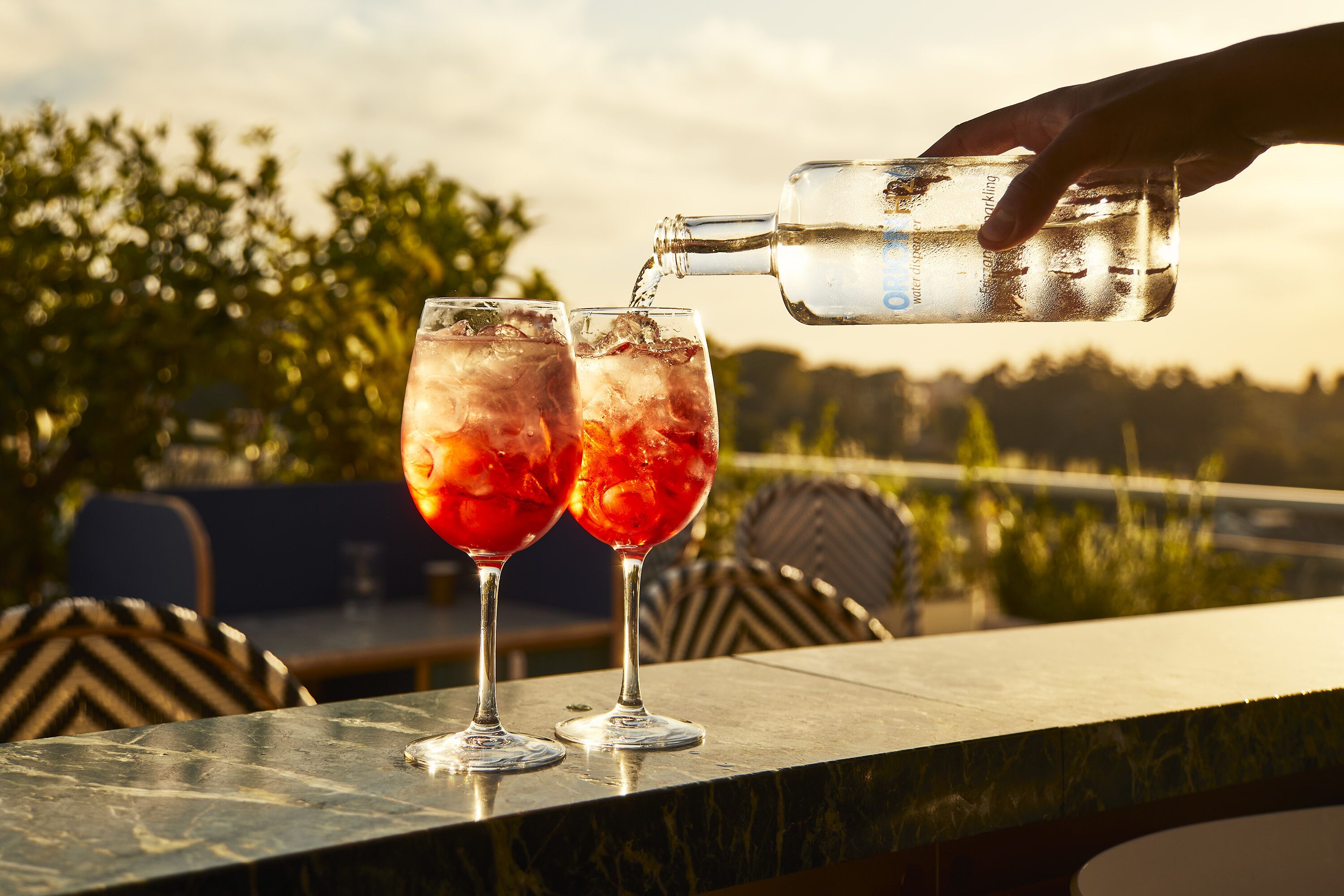 A hand pouring a clear liquid into two glasses with ice cubes on a marble countertop.