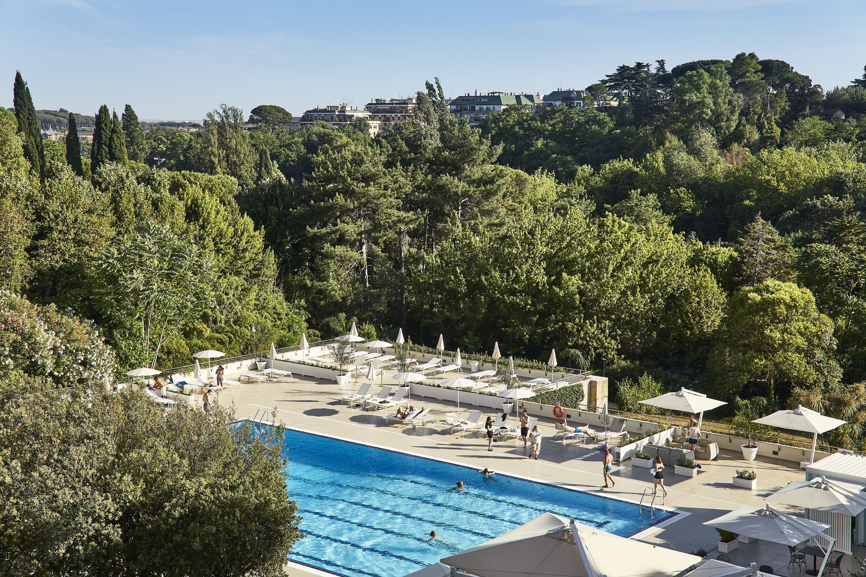 Un espace piscine avec des chaises longues et des parasols, entouré d’arbres et de bâtiments en arrière-plan.