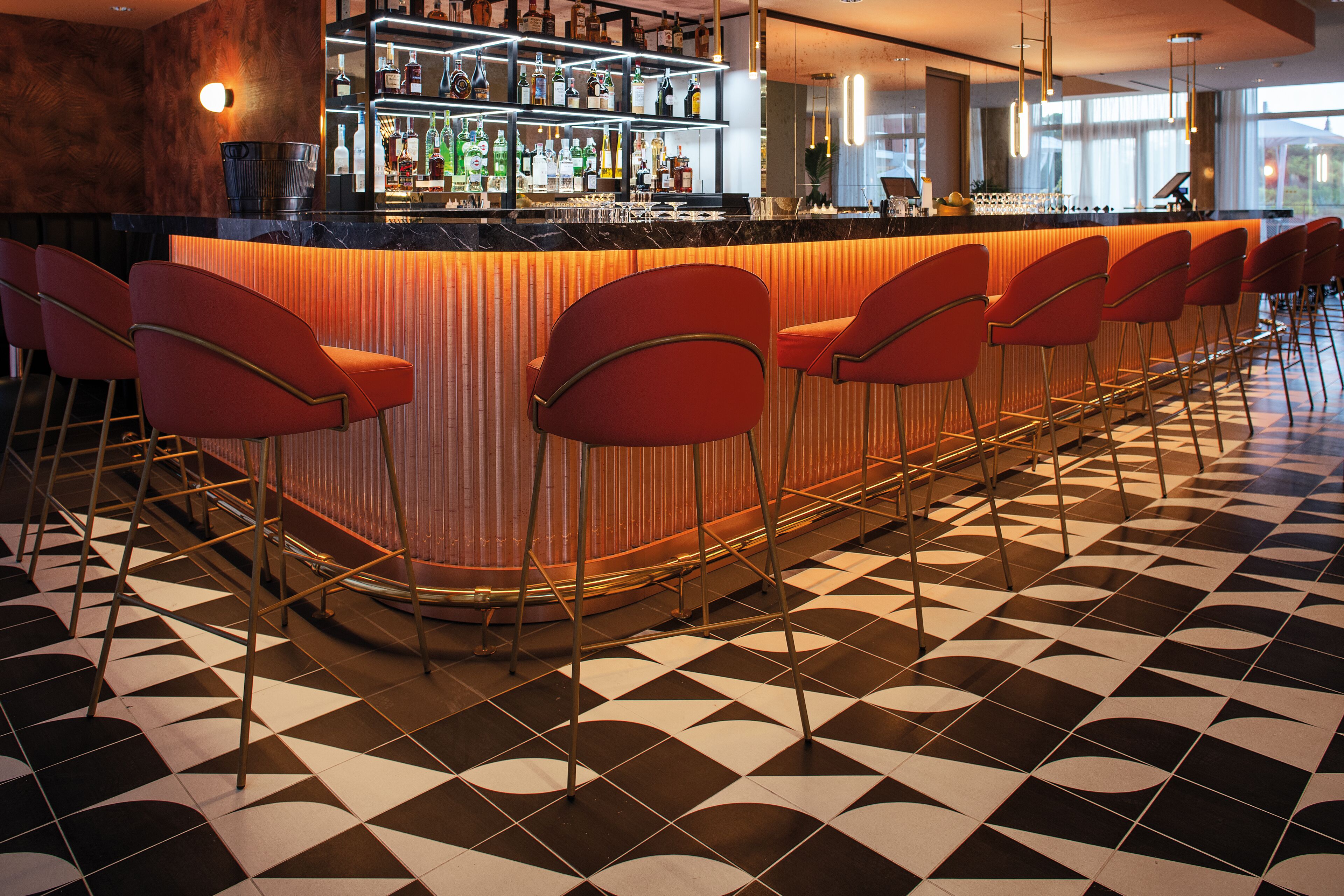 Een bar met een zwart-wit geruit vloertje, een toog en meerdere rode barkrukken.