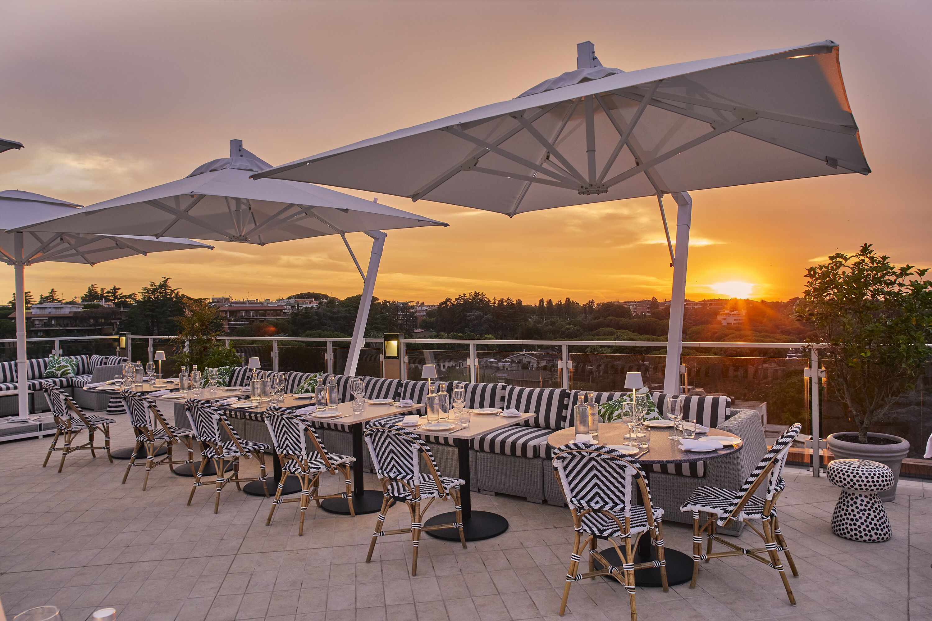 Un área de comedor al aire libre con mesas y sillas, sombrillas grandes y vista al atardecer.
