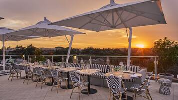 Un área de comedor al aire libre con mesas y sillas, sombrillas grandes y vista al atardecer.