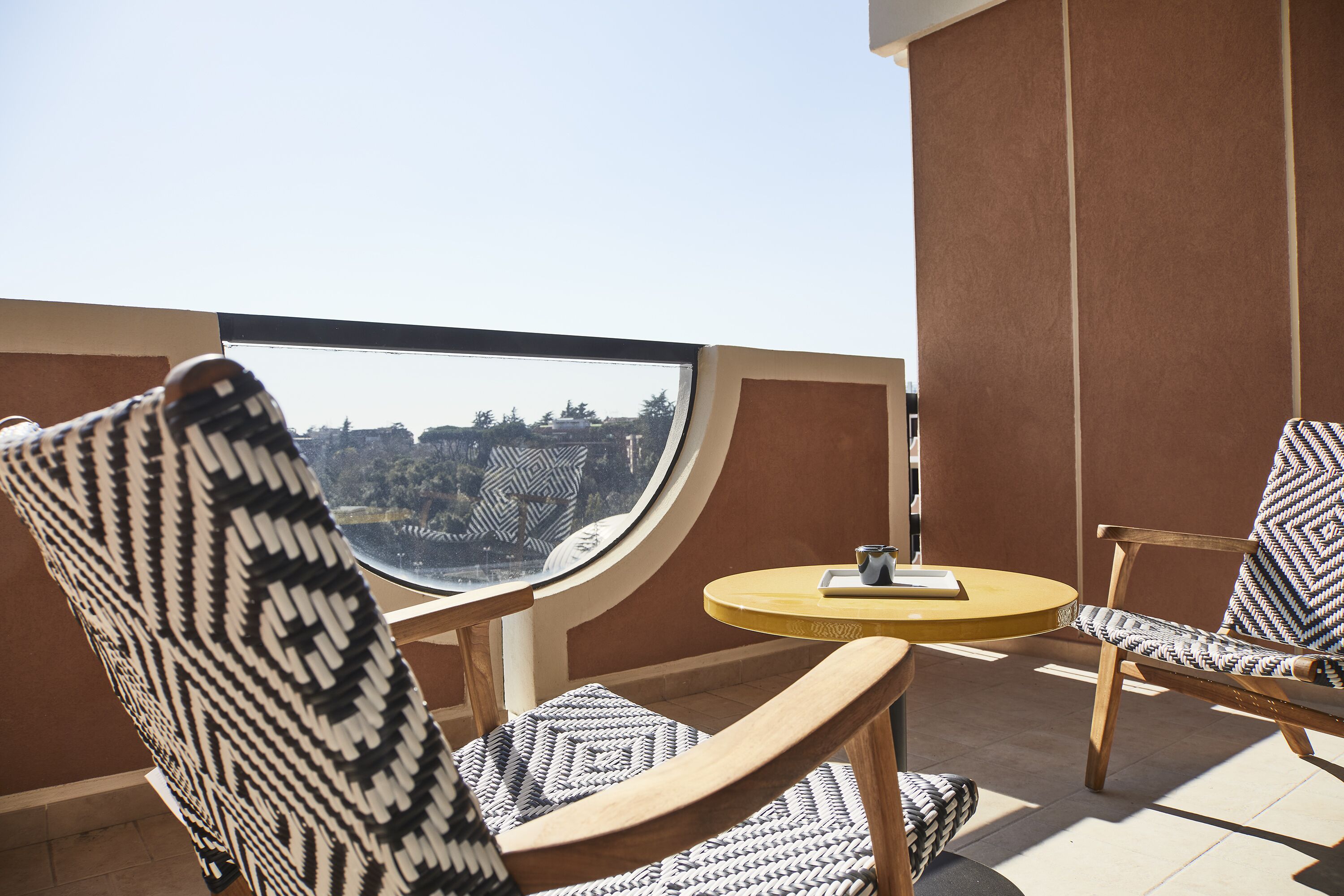 Een balkon met twee rieten stoelen en een kleine ronde tafel, met een helder uitzicht op een stadsbeeld.