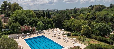 A large swimming pool with sun loungers and umbrellas, surrounded by lush greenery and trees.