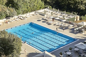 Una grande piscina all'aperto con diverse corsie, circondata da lettini e ombrelloni, e ombreggiata da alberi.