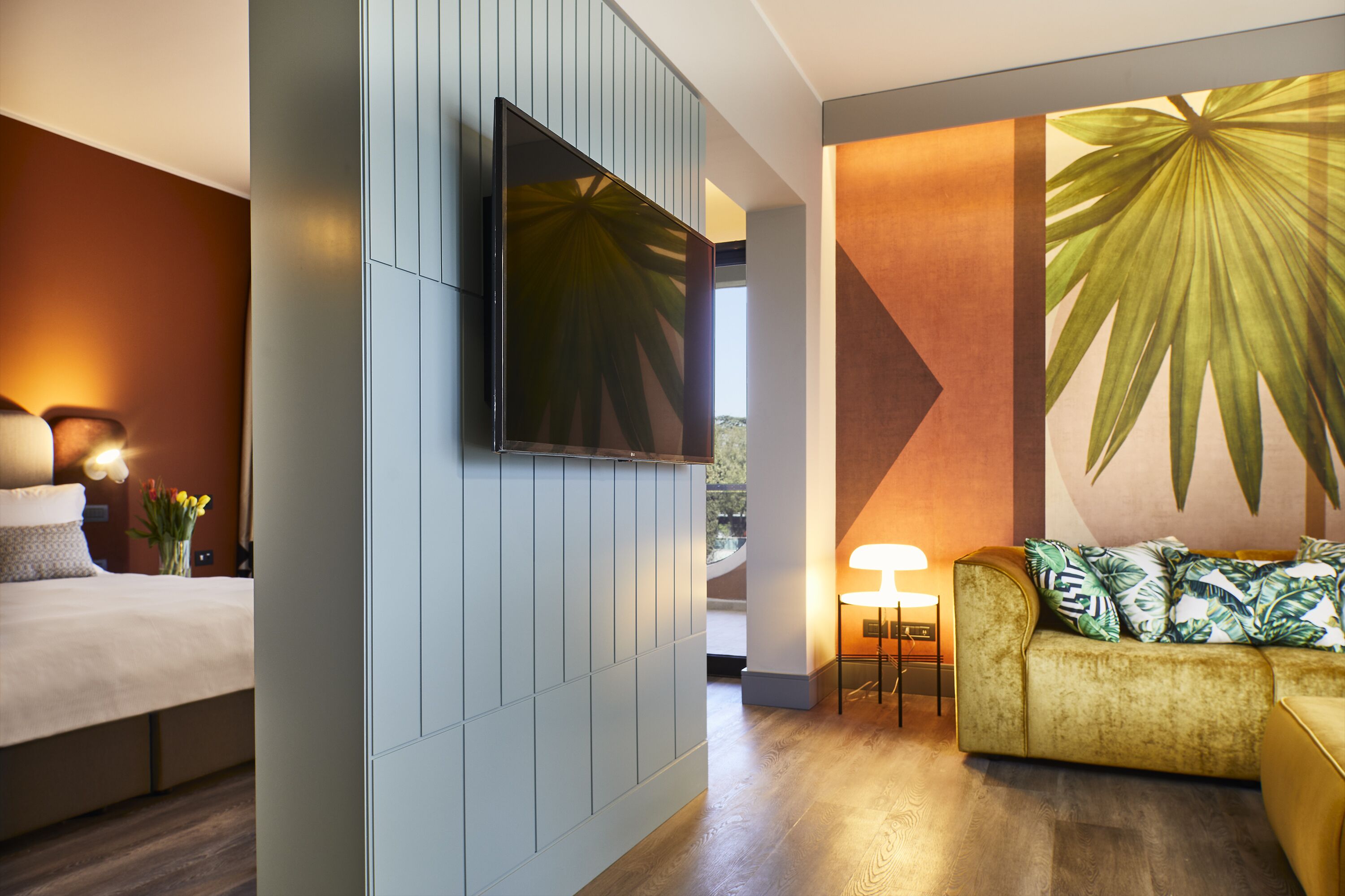 Ein modernes Hotelzimmer mit Bett, einer Couch, einem an der Wand montierten Fernseher und einem großen Wandgemälde mit tropischen Blättern.