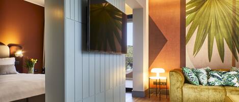 A modern hotel room with a bed, a sofa, a TV mounted on a wall, and a large tropical leaf mural.